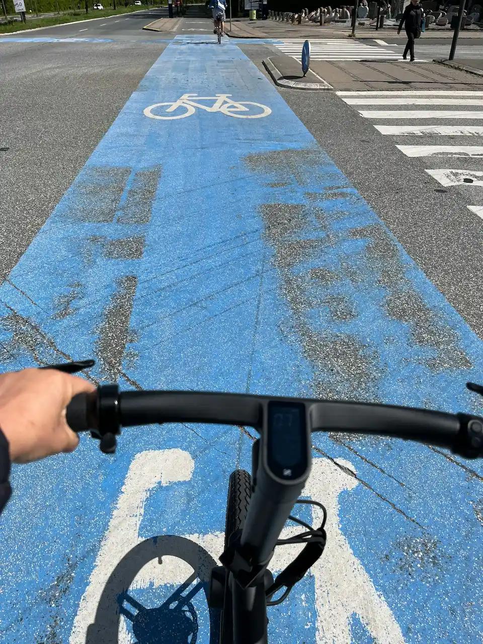 View from a cyclist riding in a designated blue bike lane, showing the handlebar and part of the bike path ahead with a bicycle symbol on the ground.