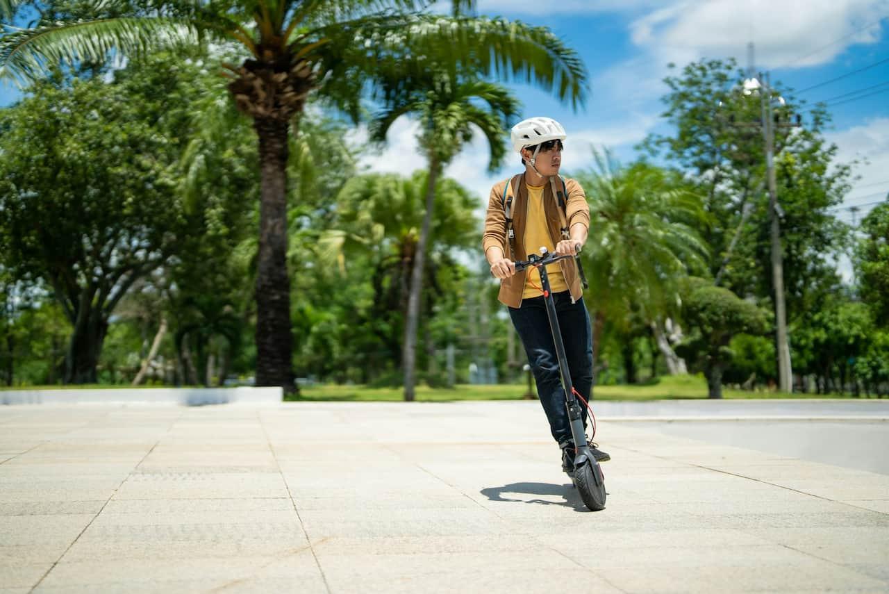 Ein Mann mit Helm fährt an einem sonnigen Tag mit einem E-Scooter durch einen Park.