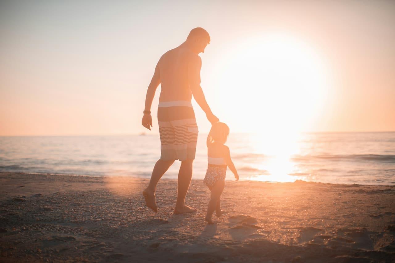 Vater und Kind spazieren bei Sonnenuntergang am Strand entlang, die Hand in Hand, während die Sonne über dem Meer untergeht.