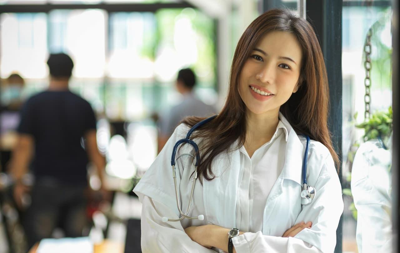 A smiling female doctor with a stethoscope around her neck, standing confidently with folded arms in a bright medical office.