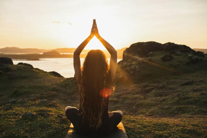 Frau in einer Yoga-Pose bei Sonnenuntergang auf einer grünen Wiese mit Blick auf Berge.